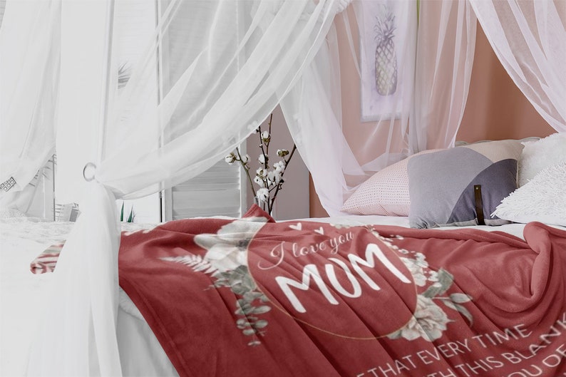 Puede incluir: Una manta de forro polar roja con un dise&ntilde;o de corona floral y el texto "I love you Mom" en un c&iacute;rculo dorado. La manta est&aacute; sobre una cama con ropa de cama blanca y un dosel.