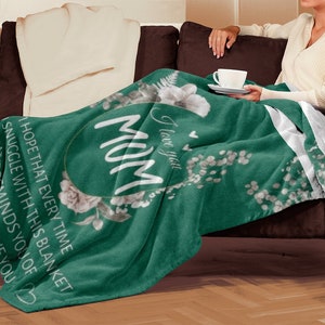 Puede incluir: Una manta de forro polar verde con un dise&ntilde;o floral blanco y el texto "Te amo Mam&aacute;" en un c&iacute;rculo.