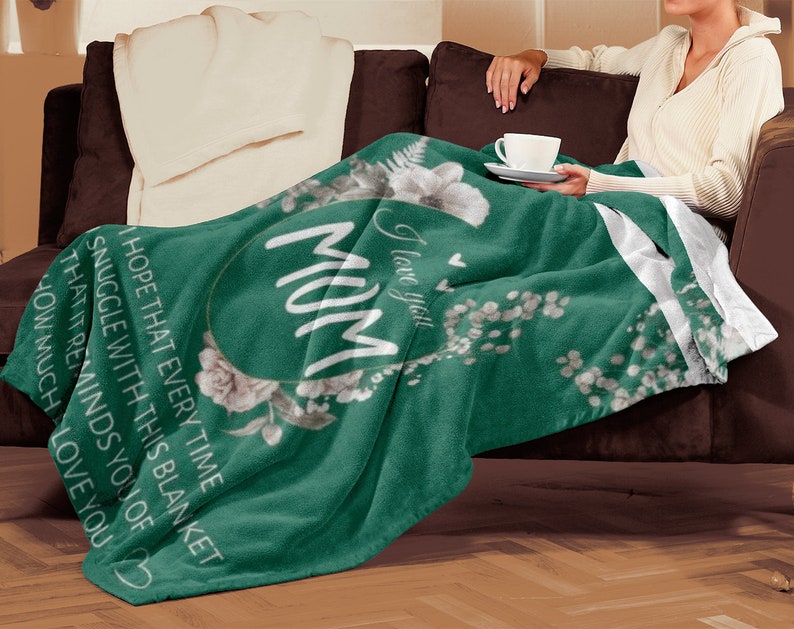 Puede incluir: Una manta de forro polar verde con un dise&ntilde;o floral blanco y el texto "Te amo Mam&aacute;" en un c&iacute;rculo.