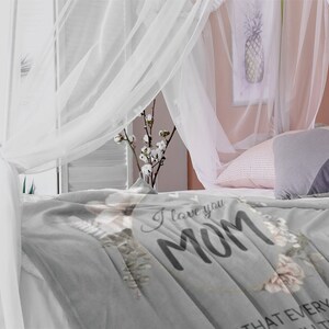 Puede incluir: Manta de franela gris con el texto "Te amo Mam&aacute;" y detalles florales. La manta est&aacute; sobre una cama con cortinas y almohadas blancas.