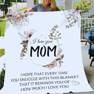 Puede incluir: Una manta blanca con un dise&ntilde;o floral y el texto "I love you MOM". El texto est&aacute; en un c&iacute;rculo dorado con un dise&ntilde;o floral. La manta tambi&eacute;n tiene el texto "I hope that every time you snuggle with this blanket that it reminds you of how much I love you".