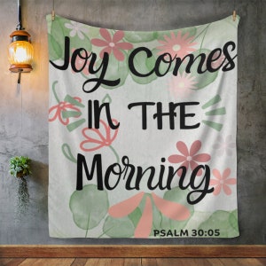 May include: A white tapestry with a watercolor floral design. The tapestry features the text "Joy Comes in the Morning" with the scripture reference "PSALM 30:05".