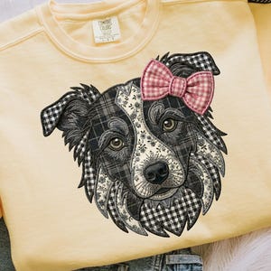 May include: A pale yellow crewneck sweatshirt featuring a large embroidered graphic of a dog's face. The dog has black and white plaid and floral patterned fur, with a pink and white gingham bow attached to its ear. The label reads Comfort Colors.