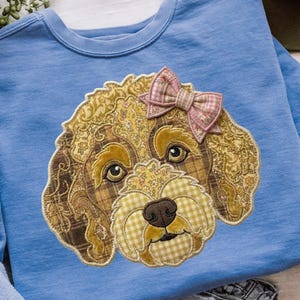 May include: A light blue crewneck sweatshirt featuring a large, central applique of a dog's face. The dog is constructed from various brown, gold, and cream fabric patches with plaid and floral patterns, and wears a pink plaid fabric bow.
