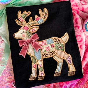 Può includere: Una t-shirt nera con una grande applicazione centrale a forma di renna. La renna è composta da vari ritagli di tessuto con motivi floreali, a quadri e leopardati, rifiniti con cuciture color cuoio e decorati con un fiocco in velluto rosa.