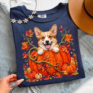May include: A navy blue t-shirt featuring a graphic of a smiling Corgi dog peeking out from a large orange pumpkin. The design is surrounded by colorful autumn leaves and vines. The shirt has a Comfort Colors brand label at the inner neckline.