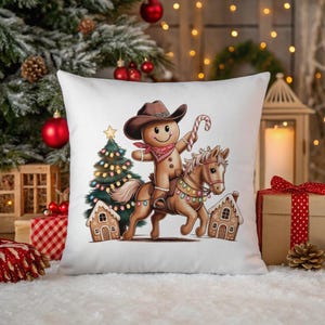 K&ouml;nnte beinhalten: Ein wei&szlig;es quadratisches Dekokissen mit einer Lebkuchenfigur, die einen braunen Cowboyhut und ein rotes Halstuch tr&auml;gt und auf einem braunen Pferd reitet. Das Motiv zeigt zudem einen geschm&uuml;ckten Weihnachtsbaum, zwei Lebkuchenh&auml;user und eine Zuckerstange.
