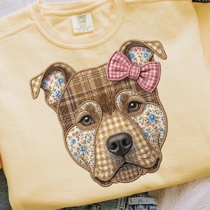 May include: A pale yellow crewneck sweatshirt featuring a large embroidered dog head graphic. The dog is composed of brown plaid and blue floral fabric patches, with a pink and white gingham bow attached to one ear. The brand label reads Comfort Colors.