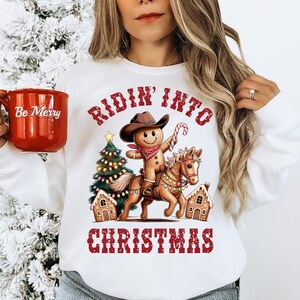 K&ouml;nnte beinhalten: Ein wei&szlig;es Sweatshirt mit Rundhalsausschnitt und einer Grafik eines Lebkuchenmanns, der einen Cowboyhut und ein Halstuch tr&auml;gt und auf einem Pferd reitet. Das Design zeigt einen Weihnachtsbaum, Lebkuchenh&auml;user und den roten Schriftzug Ridin' Into Christmas.