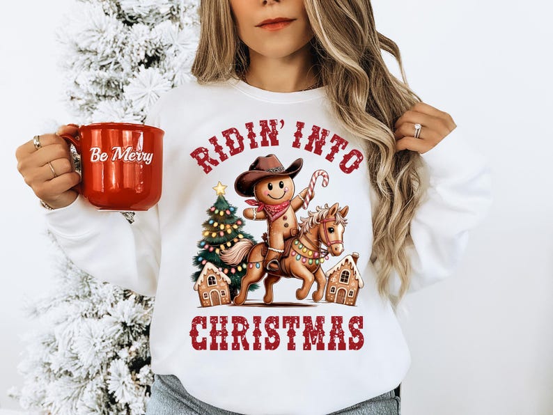 K&ouml;nnte beinhalten: Ein wei&szlig;es Sweatshirt mit Rundhalsausschnitt und einer Grafik eines Lebkuchenmanns, der einen Cowboyhut und ein Halstuch tr&auml;gt und auf einem Pferd reitet. Das Design zeigt einen Weihnachtsbaum, Lebkuchenh&auml;user und den roten Schriftzug Ridin' Into Christmas.