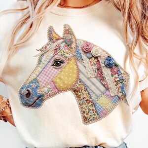 Könnte beinhalten: Ein cremefarbenes T-Shirt mit einer großen, zentralen Grafik eines Pferdekopfes. Das Pferd besteht aus verschiedenen Patchwork-Stoffmustern in Blau-, Gelb-, Rosa- und Weißtönen, akzentuiert mit kleinen Stoffblumen und Spitzenbesatz.