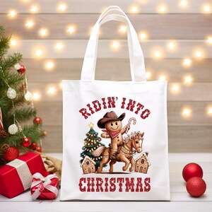 K&ouml;nnte beinhalten: Eine wei&szlig;e Tragetasche mit einer Grafik eines Lebkuchenmanns, der einen Cowboyhut und ein rotes Bandana tr&auml;gt und auf einem Pferd reitet. Das Design umfasst einen Weihnachtsbaum, zwei Lebkuchenh&auml;user und den roten Schriftzug Ridin' Into Christmas.