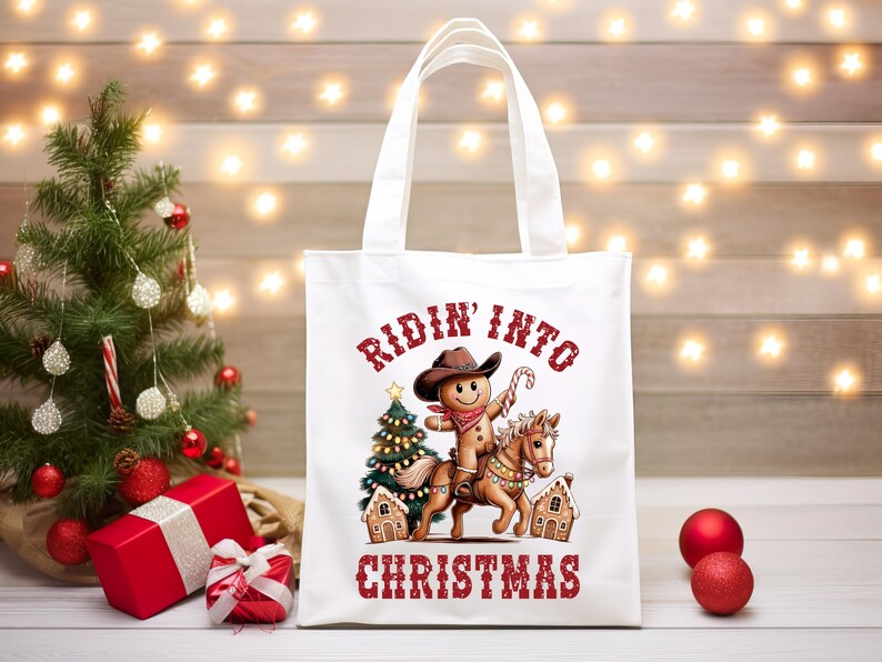 K&ouml;nnte beinhalten: Eine wei&szlig;e Tragetasche mit einer Grafik eines Lebkuchenmanns, der einen Cowboyhut und ein rotes Bandana tr&auml;gt und auf einem Pferd reitet. Das Design umfasst einen Weihnachtsbaum, zwei Lebkuchenh&auml;user und den roten Schriftzug Ridin' Into Christmas.