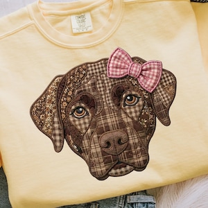 May include: A light yellow sweatshirt featuring a detailed applique of a dog's face. The dog's face is made of various brown patterned fabrics, with a pink gingham bow. The sweatshirt has long sleeves and a crew neck.