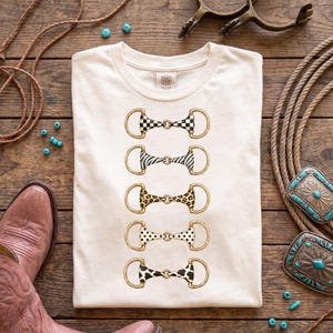 Peut inclure: Un t-shirt couleur crème présentant un motif vertical de cinq mors de cheval. Chaque mors est illustré avec un motif différent : damier noir et blanc, rayures zébrées, taches de léopard, pois noirs et imprimé vache noir et blanc.