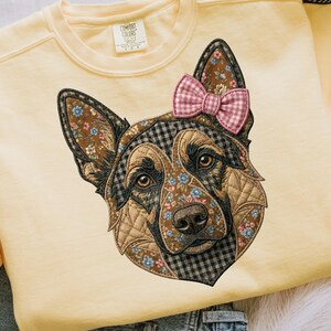 Puede incluir: Sudadera amarilla clara con un gráfico detallado de la cabeza de un pastor alemán. La cara del perro está adornada con patrones florales y a cuadros, y un lazo rosa a cuadros. La sudadera tiene cuello redondo y mangas largas.