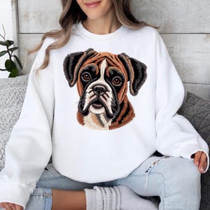 May include: A white long-sleeve sweatshirt featuring a detailed, embroidered-style graphic of a brown and black Boxer dog face centered on the chest. The dog has dark brown eyes, a black muzzle, and white markings on its forehead and snout.