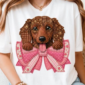 Puede incluir: Una camiseta blanca con un gráfico de un perro salchicha marrón con un pelaje estilo patchwork, que incluye patrones de cuadros y lunares marrones. El perro está colocado detrás de un gran lazo rosa con motivos florales y de cuadros.