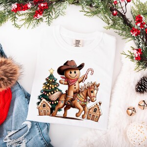K&ouml;nnte beinhalten: Ein wei&szlig;es T-Shirt mit einer Grafik eines Lebkuchenmanns, der einen braunen Cowboyhut und ein rotes Bandana tr&auml;gt und auf einem hellbraunen Pferd reitet. Das Pferd ist mit bunten Lichterketten geschm&uuml;ckt. Ein Weihnachtsbaum und Lebkuchenh&auml;user sind zu sehen.