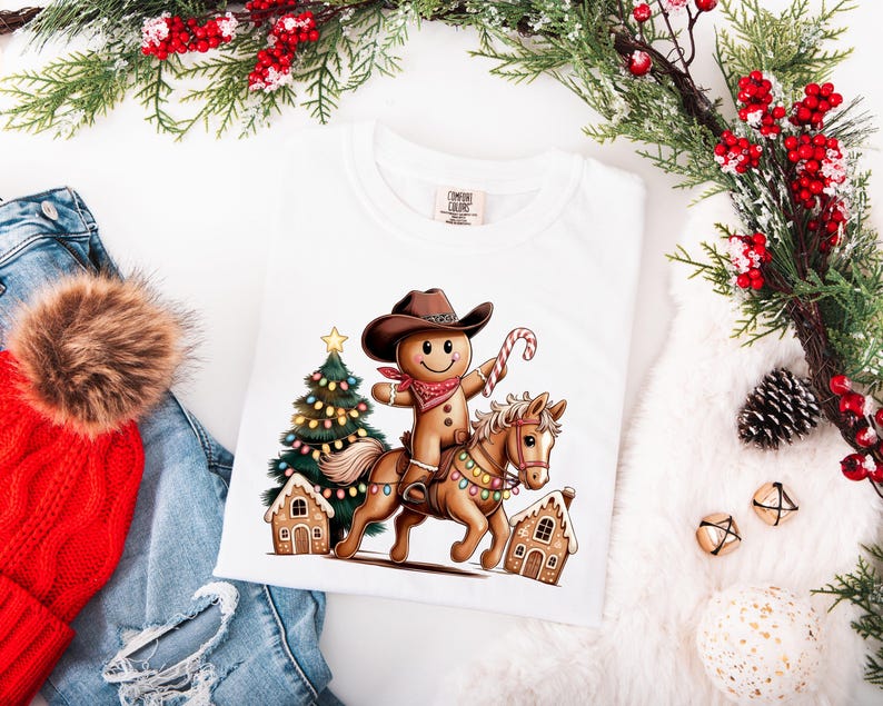 K&ouml;nnte beinhalten: Ein wei&szlig;es T-Shirt mit einer Grafik eines Lebkuchenmanns, der einen braunen Cowboyhut und ein rotes Bandana tr&auml;gt und auf einem hellbraunen Pferd reitet. Das Pferd ist mit bunten Lichterketten geschm&uuml;ckt. Ein Weihnachtsbaum und Lebkuchenh&auml;user sind zu sehen.
