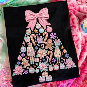 May include: A black t-shirt featuring a large, triangular Christmas tree graphic composed of various pastel-colored ornaments, including a pink bow at the top, nutcrackers, gingerbread men, candy canes, gift boxes, snowflakes, and poinsettias.