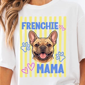 Peut inclure: Un t-shirt blanc avec un motif représentant le visage d'un bouledogue français beige centré sur des rayures verticales jaunes. Le texte "FRENCHIE MAMA" est imprimé en bleu, avec des cœurs roses et des empreintes de pattes bleues.