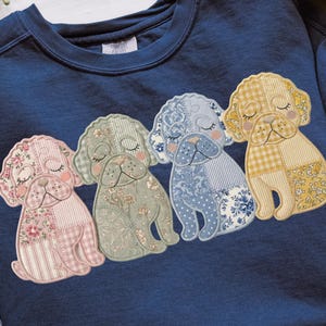 May include: A navy blue sweatshirt with four appliqued dog designs. Each dog is made from patchwork fabrics in pastel shades, including pink floral, green floral, blue floral, and yellow gingham. The dogs have closed eyes and rosy cheeks.