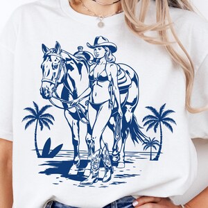 以下が含まれることがあります： 白い半袖Tシャツ。ビキニ、カウボーイハット、ブーツを身につけた人物が、サドルをつけた馬と一緒にヤシの木のあるビーチを歩いている様子が、濃い青色のグラフィックでプリントされています。細部まで描き込まれたイラストです。