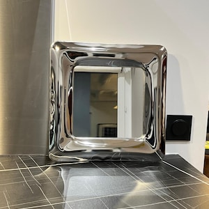 以下が含まれることがあります： 厚みがあり、わずかに波打ったフレームを持つ正方形の銀色の鏡。鏡は黒いタイル張りの表面の上に置かれています。