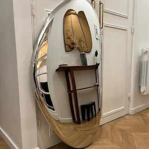 May include: A large, oval-shaped floor mirror with a polished silver frame. The mirror has a convex, curved surface that creates a distorted, wide-angle reflection of the surrounding room, including a wooden console table and a small black stool.