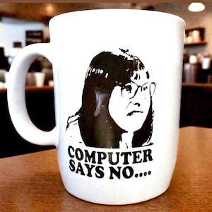 Op de afbeelding: Witte keramische mok met een zwart-wit afbeelding van een vrouw met een bril en de tekst "COMPUTER SAYS NO...." eronder. De mok heeft een gebogen handvat. De mok staat op een houten oppervlak.