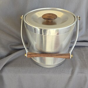 May include: A vintage ice bucket with a silver-colored, ribbed exterior and a matching lid. The lid features a round, wooden knob. The bucket has a handle and a wooden bar handle. A classic barware item.