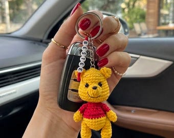 Op de afbeelding: Een gehaakte sleutelhanger met een geel en rood berenfiguurtje bevestigd aan een zilveren metalen ring. Een klein wit kralensnoer met zwarte letters die het woord BESTEE spellen, is aan de ring boven het berenfiguurtje bevestigd.