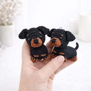 Op de afbeelding: Twee handgemaakte gehaakte teckelpuppy's. De honden zijn zwart met bruine accenten op hun snuit, poten en oren. Ze hebben kleine, zwarte ogen en zitten rechtop. De puppy's zijn ongeveer 8 cm hoog.