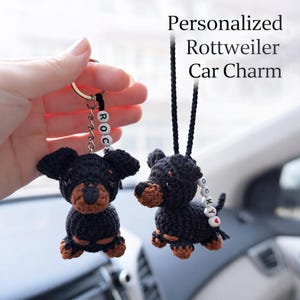 Op de afbeelding: Twee zwart-bruine gehaakte Rottweiler auto-bedels. E&eacute;n is bevestigd aan een sleutelhanger met de letters "ROC". De andere hangt aan een zwart koord. De tekst "Personalized Rottweiler Car Charm" staat op de achtergrond.