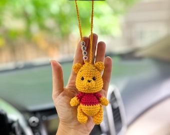 Op de afbeelding: Een handgemaakte gele en rode gehaakte Winnie de Poeh auto-hanger. De hanger hangt aan een gele draad met de naam "JENNY" gespeld in kralen. De hanger wordt voor het interieur van een auto gehouden.