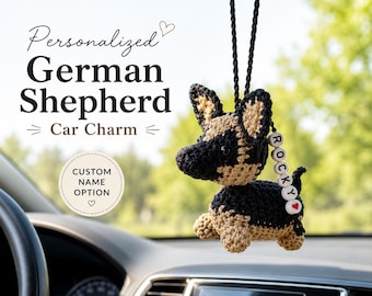 Op de afbeelding: Een gehaakte auto-hanger in de vorm van een Duitse herder, hangend aan een zwart koord. De figuur van zwart en beige garen heeft donkere ogen en een klein wit kralensnoer met de naam ROCKY en een rood hartje eraan bevestigd.