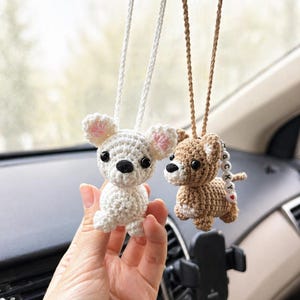 Op de afbeelding: Twee gehaakte hondenornamenten die aan de achteruitkijkspiegel van een auto hangen. E&eacute;n is wit met roze accenten, de andere is bruin. Beide hebben zwarte ogen en hangen aan koorden.