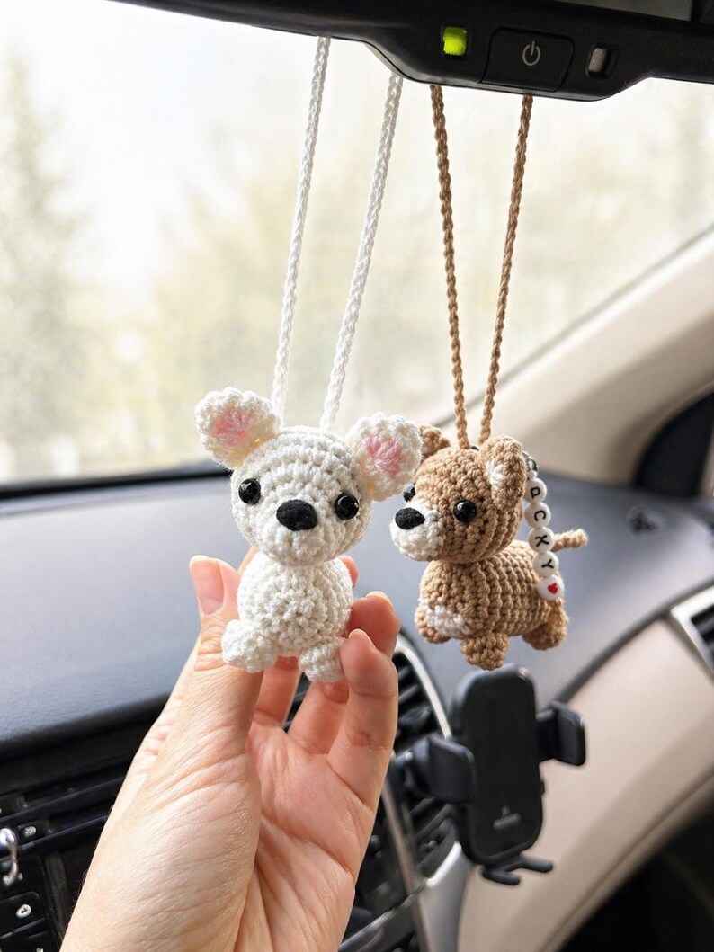 Op de afbeelding: Twee gehaakte hondenornamenten die aan de achteruitkijkspiegel van een auto hangen. E&eacute;n is wit met roze accenten, de andere is bruin. Beide hebben zwarte ogen en hangen aan koorden.