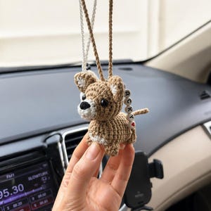 Op de afbeelding: Gehaakte bruine hondenauto-hanger met witte accenten en een klein rood hart. De hanger hangt aan een bruin en zilverkleurig touwtje. De hond heeft een kleine staart met de letters "RCK".