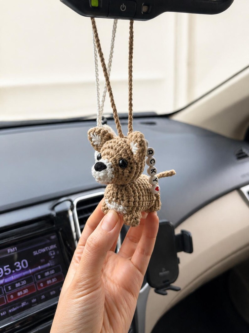 Op de afbeelding: Gehaakte bruine hondenauto-hanger met witte accenten en een klein rood hart. De hanger hangt aan een bruin en zilverkleurig touwtje. De hond heeft een kleine staart met de letters "RCK".