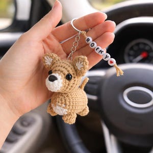 Op de afbeelding: Gehaakte bruine honden sleutelhanger met witte accenten. De hond heeft zwarte ogen en een zwarte neus. De sleutelhanger bevat een kralenreeks met de tekst "ROCKY". Een kleine rode hartkralen is ook inbegrepen.