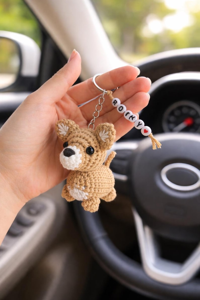 Op de afbeelding: Gehaakte bruine honden sleutelhanger met witte accenten. De hond heeft zwarte ogen en een zwarte neus. De sleutelhanger bevat een kralenreeks met de tekst "ROCKY". Een kleine rode hartkralen is ook inbegrepen.
