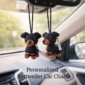Op de afbeelding: Twee zwart-bruine Rottweiler auto-bedels die aan de achteruitkijkspiegel van een auto hangen. De gehaakte bedels hebben een gepersonaliseerd naamlabel. De tekst onderaan luidt "Personalized Rottweiler Car Charm."