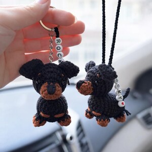 Op de afbeelding: Twee gehaakte zwart-bruine hondjes als ornament. De ene is een sleutelhanger, de andere hangt aan een zwart koord. Beide hebben witte kralen met de naam ROCKY en een rood hartje. De hondjes hebben zwarte lichamen met bruine snuiten en pootjes.