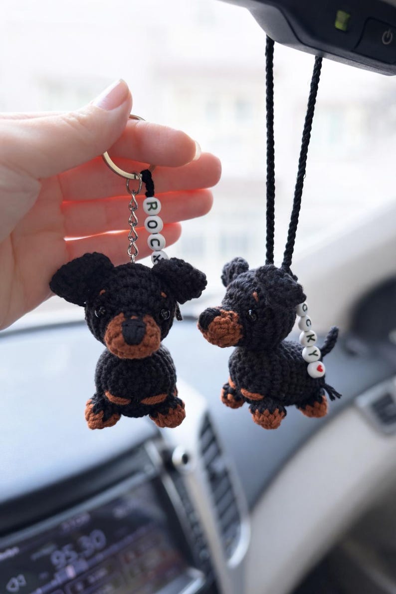 Op de afbeelding: Twee gehaakte zwart-bruine hondjes als ornament. De ene is een sleutelhanger, de andere hangt aan een zwart koord. Beide hebben witte kralen met de naam ROCKY en een rood hartje. De hondjes hebben zwarte lichamen met bruine snuiten en pootjes.