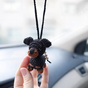 Op de afbeelding: Handgemaakte gehaakte hondenauto-hanger in zwart en bruin. De kleine hond heeft een zwart lichaam, een bruine snuit en poten. Hij hangt aan een zwart koord, ideaal voor de achteruitkijkspiegel.