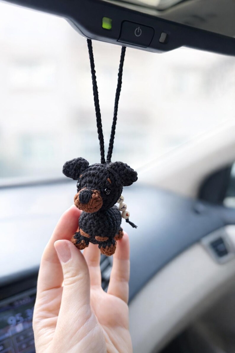 Op de afbeelding: Handgemaakte gehaakte hondenauto-hanger in zwart en bruin. De kleine hond heeft een zwart lichaam, een bruine snuit en poten. Hij hangt aan een zwart koord, ideaal voor de achteruitkijkspiegel.