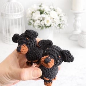 Op de afbeelding: Twee kleine gehaakte teckelbeeldjes gemaakt van zwart en bruin garen. Elke hond heeft hangende oren, een afgeronde snuit met bruine accenten en kleine zwarte kraaloogjes. De figuren zijn gemaakt met een gestructureerd steekpatroon.