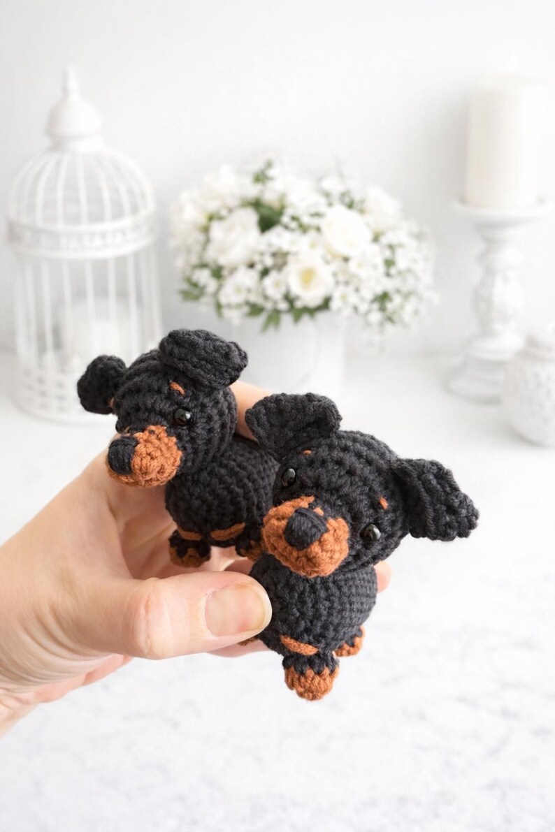 Op de afbeelding: Twee kleine gehaakte teckelbeeldjes gemaakt van zwart en bruin garen. Elke hond heeft hangende oren, een afgeronde snuit met bruine accenten en kleine zwarte kraaloogjes. De figuren zijn gemaakt met een gestructureerd steekpatroon.